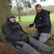 Helping a Victim at First Aid Workshop Two men enjoy an adventurous workshop outdoors in Utrecht. One man sits laughing on a green foam cushion against a tree, dressed in winter clothing. The other kneels beside him and gives a thumbs up. Join this unique outdoor first aid course!