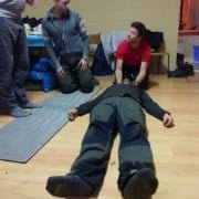 Instruction at the First Aid Workshop Experience an adventurous first aid workshop in nature! Three participants practice life-saving procedures on a wooden floor, surrounded by blue mats and benches. The realistic setting with props teaches you to react quickly. Escape the routine and learn essential rescue skills for every adventurer. Join us and be prepared!