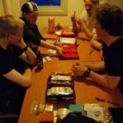 Review Expedition First Aid Kit A group of four people sits comfortably around a wooden table, filled with camping gear and first aid kits. With drinks within reach, they laugh and chat. They're in a cozy cabin in Utrecht with a nighttime scene outside the window. Ready for adventure? Join us!