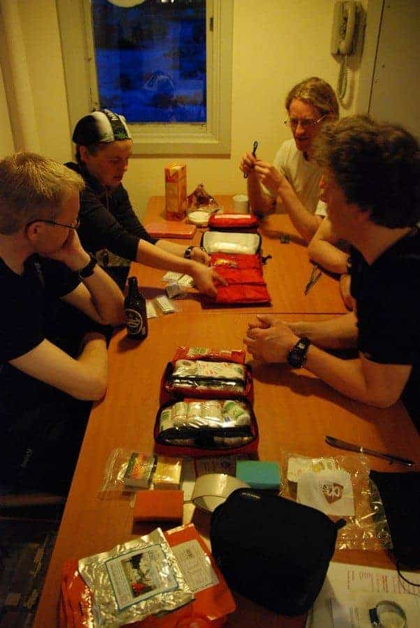 Review Expedition First Aid Kit A group of four people sits comfortably around a wooden table, filled with camping gear and first aid kits. With drinks within reach, they laugh and chat. They're in a cozy cabin in Utrecht with a nighttime scene outside the window. Ready for adventure? Join us!
