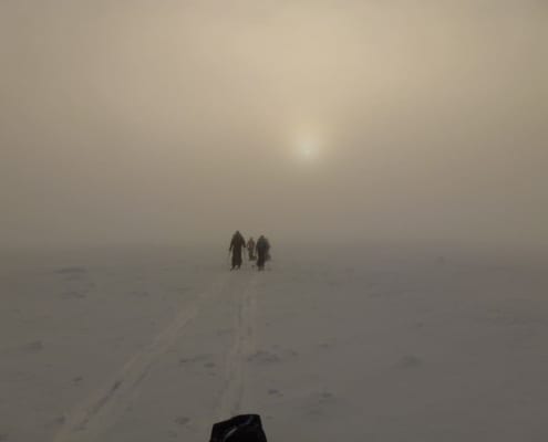 Three people walk in the snow towards a vague, distant light, leaving tracks in the misty landscape.