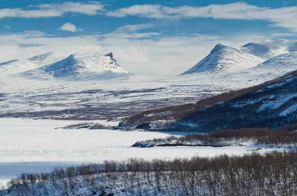 Snow-covered landscape with distant mountains, including the iconic Lapporten, under a blue sky with scattered clouds. In the foreground lies a largely frozen body of water, partially surrounded by leafless trees. 