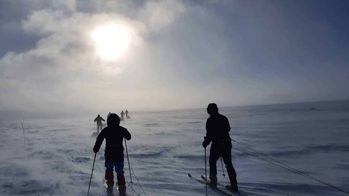 Verschillende mensen skiën in een besneeuwd, winderig landschap met slecht zicht en de zon schijnt door een bewolkte hemel.