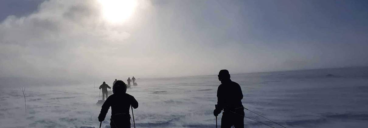 Verschillende mensen skiën in een besneeuwd, winderig landschap met slecht zicht en de zon schijnt door een bewolkte hemel.