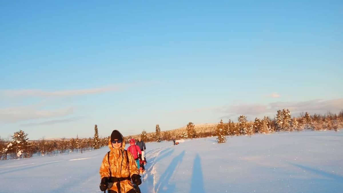 Mensen lopen in een besneeuwd landschap en trekken sleeën met proviand onder een strakblauwe lucht.
