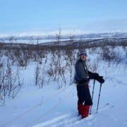 Een persoon in winteruitrusting staat in een besneeuwd landschap met schaarse bomen, houdt skistokken vast en is met een touw aan een slee vastgemaakt. Op de achtergrond zijn met sneeuw bedekte heuvels zichtbaar onder een helderblauwe lucht.