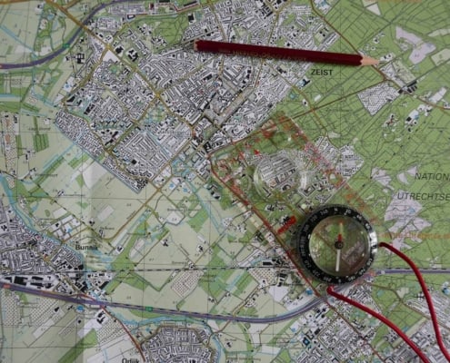 Een kleurrijke topografische kaart van de Utrechtse Heuvelrug, met groene en beige gebieden die verschillende terreinen tonen. Een transparante kompas op de kaart wijst de richting aan. Een rood potlood ligt diagonaal over het midden van de kaart, waar ook stedelijke gebieden en locaties gemarkeerd zijn. Perfect voor het volgende jouw avontuur!