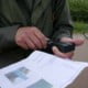 Frank Moison on the Navigating with Map and Compass Workshop in the Netherlands 2 An adventurer in a green jacket stands outside navigating with a GPS device in hand, while holding a map full of images. In the background, someone cycles past and you can see a green landscape in the distance.