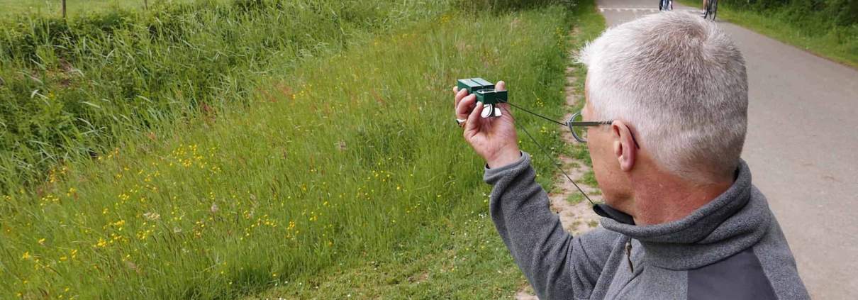 Nico Van Vondelen about the Navigating with Map and Compass Workshop in the Netherlands 1 A man with gray hair and glasses stands along a paved path, pointing his camera at grazing cows in a green field. Wild flowers and tall grass adorn the path, while trees form the background. In front of him, someone is cycling, possibly with a map for navigation.Looking for adventure? Discover beautiful cycling routes, green meadows, and enchanting landscapes! Feel free in nature and capture moments!