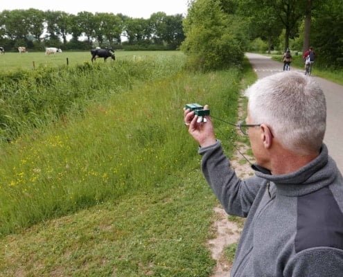 Nico Van Vondelen about the Navigating with Map and Compass Workshop in the Netherlands 11 Een man met grijs haar en bril staat langs een verhard pad en richt zijn camera op grazende koeien in een groen veld. Wilde bloemen en hoog gras sieren het pad, terwijl bomen de achtergrondvormen. Voor hem fietst iemand, mogelijk met een kaart voor navigatie.Op zoek naar avontuur? Ontdek prachtige fietsroutes, groene weides en betoverende landschappen! Voel je vrij in de natuur en leg momenten vast!