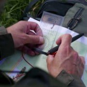 Determining location on the map with a map angle meter Discover the ultimate adventure experience! A person in a green jacket holds a compass and pen over a topographic map. In the background, a green backpack lies on the grass – perfect for map reading and outdoor discoveries. Prepare for your next adventure!