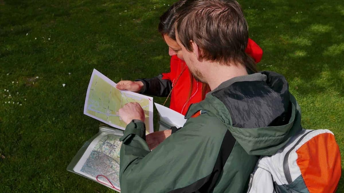Twee avonturiers staan buiten op gras, verdiept in een topografische kaart. De eerste, met licht haar en een groene jas, draagt een grijs-oranje rugzak. De tweede, met donker haar en een rode jas, zichtbaar betrouwbaar de kaart. Plan je volgende avontuur met ons!