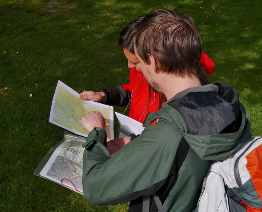 Twee avonturiers staan buiten op gras, verdiept in een topografische kaart. De eerste, met licht haar en een groene jas, draagt een grijs-oranje rugzak. De tweede, met donker haar en een rode jas, zichtbaar betrouwbaar de kaart. Plan je volgende avontuur met ons!