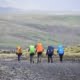Simon Van de Rivier over de Vloer is Lava Trekking in IJsland 2 Vijf avonturiers met kleurrijke rugzakken (oranje, groen, blauw) wandelen over een grindpad door een ruig, heuvelachtig landschap. De bewolkte lucht geeft de omgeving een grijze tint. Volg hun uitdagende trektocht door uitgestrekte en golvende terreinen voor de ultieme outdoor ervaring.