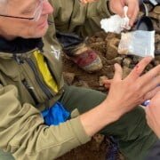 Bandaging a Cut on a Finger An older man with gray hair and glasses, dressed in a green jacket, sits on rocky ground. During a first aid workshop in Utrecht, he carefully tends to a cut on his hand. Next to him lies a supply container with extra materials.Are you ready for adventure? Book your outdoor activity in Utrecht now!