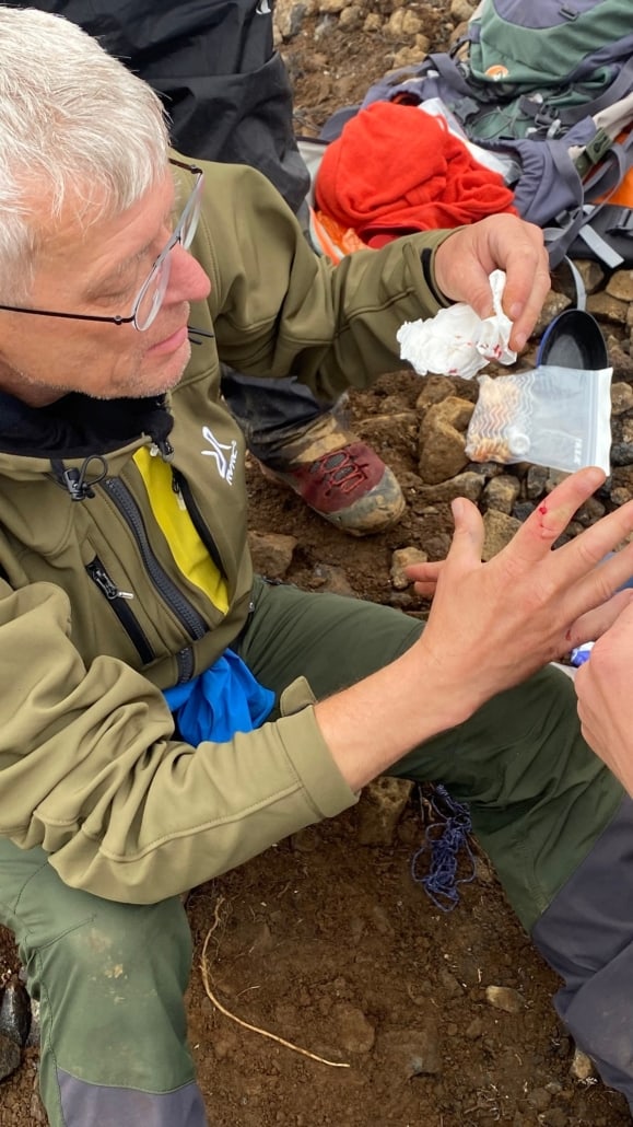 Bandaging a Cut on a Finger An older man with gray hair and glasses, dressed in a green jacket, sits on rocky ground. During a first aid workshop in Utrecht, he carefully tends to a cut on his hand. Next to him lies a supply container with extra materials.Are you ready for adventure? Book your outdoor activity in Utrecht now!