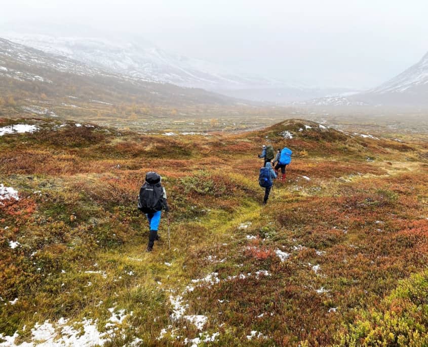 Vier avonturen met rugzakken door het uitgestrekte grasland van Sarek, Zweden. Sneeuwplekken en herfstkleurige struiken kleuren het landschap. Mist verbergt de verre bergen en geeft een serene en avontuurlijke sfeer. De groep ontdekt een vaag pad door het ruige terrein, klaar voor hun volgende avontuur.Boek nu je lengte reis naar Sarek!
