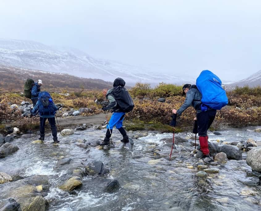 Vier avontuurlijke wandelaars, uitgerust met grote rugzakken en waterdichte kleding, steken een ondiepe, rotsachtige beek over in het mistige Sarek, Zweden. Ze banen zich een weg door nat grasland en hebben een uitzicht op besneeuwde bergtoppen. Met behulp van trekkingstokken trotseren ze de oneffen ondergrond en het water.
