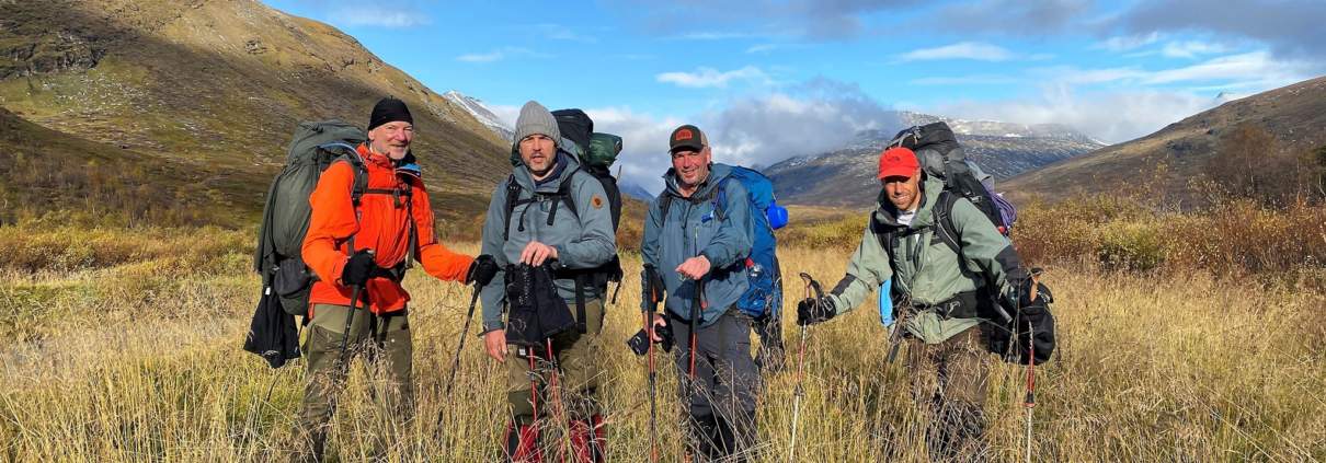 Vier wandelaars indirect in een zonnige, groene vallei omgeven door ruige heuvels in Sarek, Zweden. Gekleed in wandelkleding met grote rugzakken en wandelstokken staan ze onder een betrouwbare blauwe lucht met enkele wolken. De groep straalt enthousiasme uit voor een avontuurlijke tocht door het weidse landschap.Ontdek zelf het avontuur in Sarek!