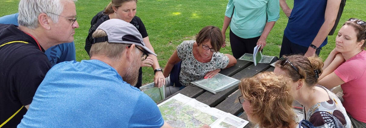 Een groep avonturiers opvallend zich rond een picknicktafel in een park in Utrecht voor een workshop kaart en kompasnavigatie. Enthousiast besproken ze de route terwijl iemand aanduidingen op de kaart maakt. Omringd door groen, klaar om het avontuur tegemoet te gaan.