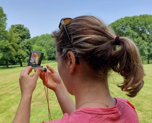 Viola about the Navigating with map and compass workshop in the Netherlands 2 Avontuurlijk bezig in Utrecht: Een persoon met een paardenstaart en roze shirt staat in een groen park. Met een klein spiegeltje en zonnebril op het hoofd, lijkt het ook alsof ze oefenen met kaart en kompas tijdens een spannende workshop. Ontdek Utrecht op soepel wijze!