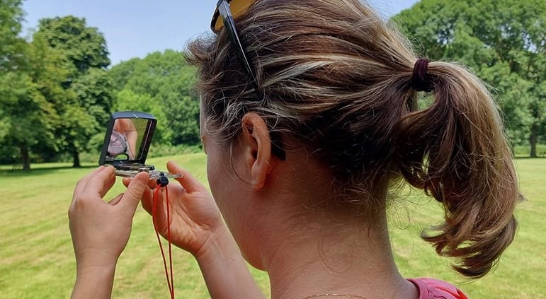 Viola about the Navigating with map and compass workshop in the Netherlands 1 Avontuurlijk bezig in Utrecht: Een persoon met een paardenstaart en roze shirt staat in een groen park. Met een klein spiegeltje en zonnebril op het hoofd, lijkt het ook alsof ze oefenen met kaart en kompas tijdens een spannende workshop. Ontdek Utrecht op soepel wijze!
