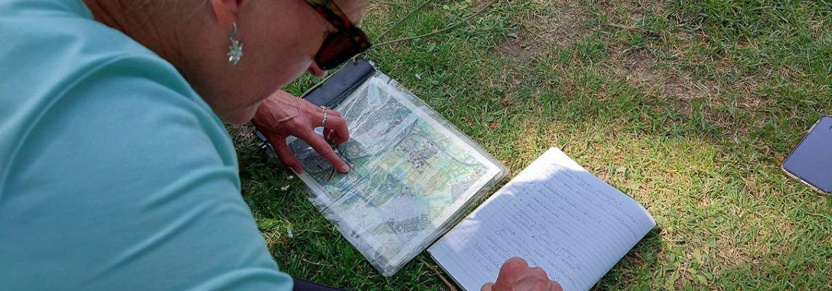 Een avontuurlijke wacht op jou! Een persoon met grijs haar, zonnebril en groen shirt bestudeert een transparante kaart terwijl hij een notitieboekje vasthoudt. Naast hem ligt een kompas en een telefoon in het gras. Ontdek de natuur; leer navigeren en verken de wildernis!