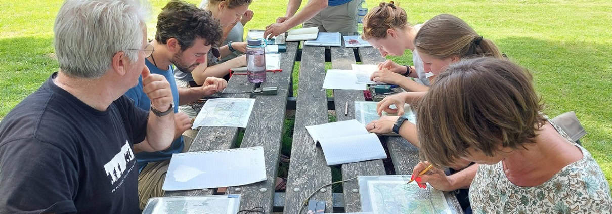 Een groep van acht mensen zit aan een picknicktafel in de natuur. Ze zijn actieve bezig met kaartlezen en schrijven. Notitieboeken, kaarten en pennen liggen verspreid op de tafel. De groene omgeving met bomen op de achtergrond wijst op een informele kaart-workshop of trainingssessie met uitleg.Boek nu deze avontuurlijke kaartleessessie en leer navigeren als een pro te midden van de natuur!