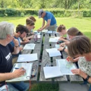 Kaart en kompas navigatie workshop_23 Een groep van acht mensen zit aan een picknicktafel in de natuur. Ze zijn actieve bezig met kaartlezen en schrijven. Notitieboeken, kaarten en pennen liggen verspreid op de tafel. De groene omgeving met bomen op de achtergrond wijst op een informele kaart-workshop of trainingssessie met uitleg.Boek nu deze avontuurlijke kaartleessessie en leer navigeren als een pro te midden van de natuur!