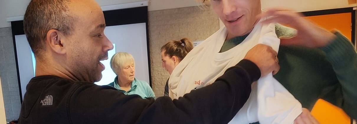 Twee mensen oefenen het gebruik van een armband tijdens een Outdoor Eerste Hulp Workshop. Op de achtergrond zijn drie bezig met anderen activiteiten en discussies. De ruimte heeft een projectiescherm en oranje accenten aan de muren. Ideaal voor avonturiers die vooraf op alles willen zijn!