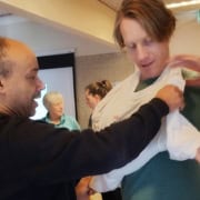 Twee mannen bereiden zich voor op een avontuur: de ene helpt de ander een mitella om te doen. Achter hen maakt een grijze vrouw en iemand met vastgebonden haar zich klaar voor een outdoor EHBO-workshop in Utrecht. Zoek je avontuur? Doe mee!