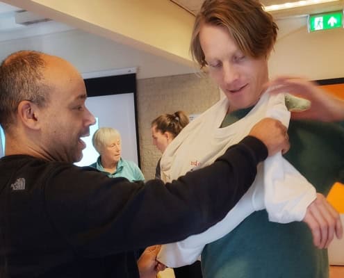 Arnaud De Wilde on the Outdoor First Aid Essentials in the Netherlands 6 Twee mensen oefenen het gebruik van een armband tijdens een Outdoor Eerste Hulp Workshop. Op de achtergrond zijn drie bezig met anderen activiteiten en discussies. De ruimte heeft een projectiescherm en oranje accenten aan de muren. Ideaal voor avonturiers die vooraf op alles willen zijn!