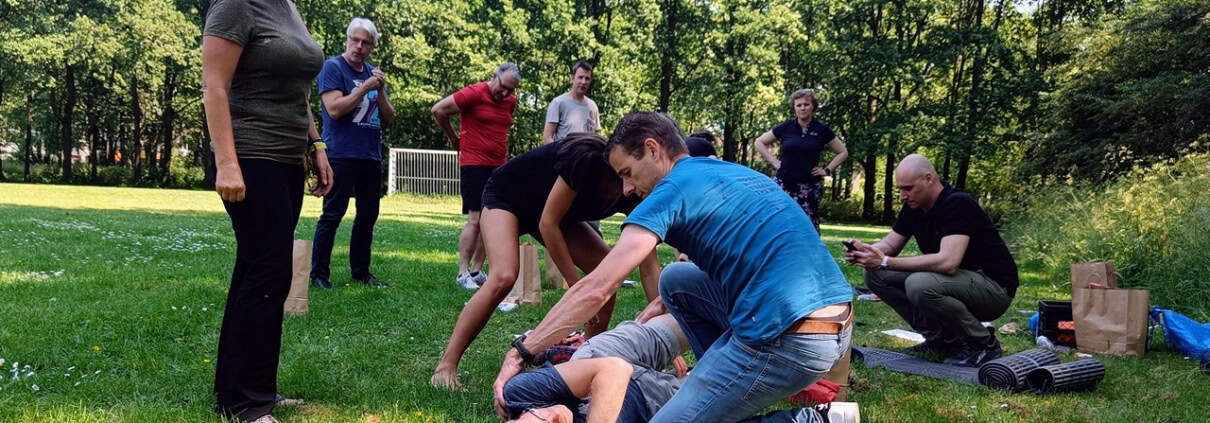 Studio Baak on the Outdoor First Aid Essentials in the Netherlands 1 Een groep mensen traint zelfverdedigingstechnieken in de buitenlucht. Op de voorgrond demonstreert een instructeur een manoeuvre op een cursist die op de grond ligt. Dit lijkt op een Outdoor EHBO-workshop waar volwassenen actief meedoen en toekijken. Omringd door bomen en dicht bij een voetbaldoel, beleef je hier avontuur en leer je essentiële vaardigheden in de frisse buitenlucht.