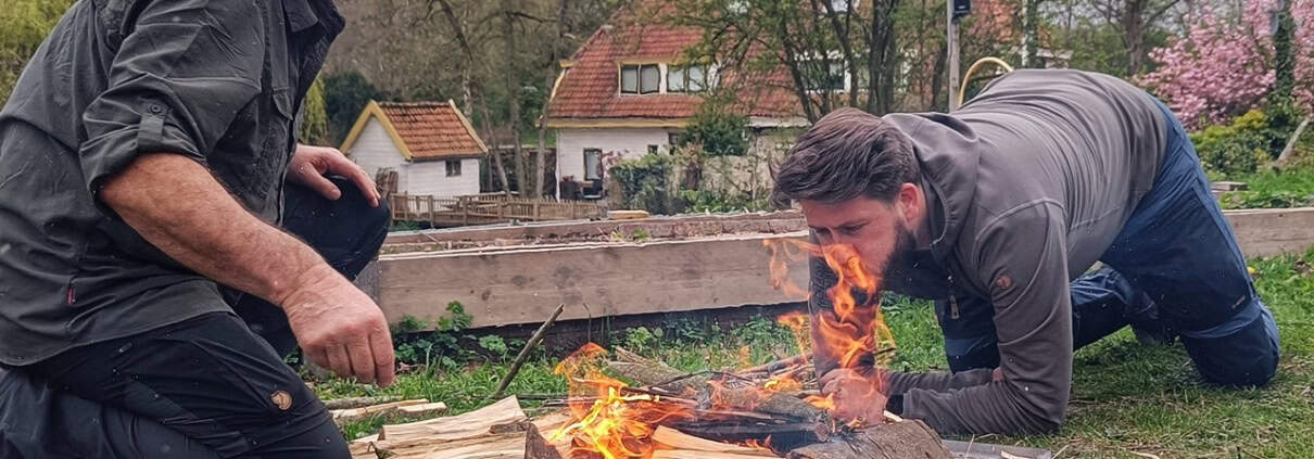 Twee mannen zijn bezig met een klein kampvuur. De een gebruikt zijn jas als windscherm, terwijl de andere het vuur aanwakkert. Omgeven door tuinen en bomen, bij een huis op de achtergrond, voelt het als een spontane houthak- en kampvuurworkshop. Perfect voor avonturiers die van buitenactiviteiten houden!