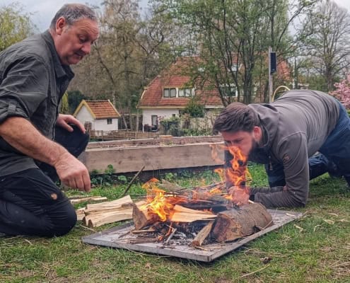 Ires Ghielen on the Lumbering and Campfire Workshop in the Netherlands 9 Twee mannen zijn bezig met een klein kampvuur. De een gebruikt zijn jas als windscherm, terwijl de andere het vuur aanwakkert. Omgeven door tuinen en bomen, bij een huis op de achtergrond, voelt het als een spontane houthak- en kampvuurworkshop. Perfect voor avonturiers die van buitenactiviteiten houden!