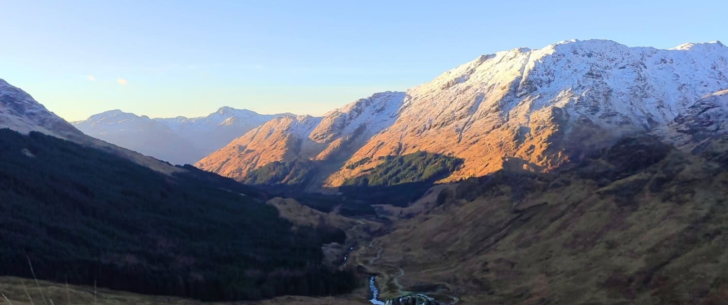 Ademend landschap met bergen en valleien in de late middagzon, met besneeuwde toppen en een rustig stroompje.