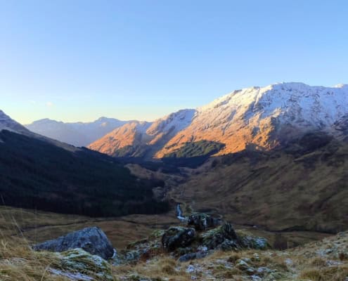 Ademend landschap met bergen en valleien in de late middagzon, met besneeuwde toppen en een rustig stroompje.