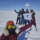 Group of adventurers poses in the snow with a wind turbine and flag in the Arctic landscape.