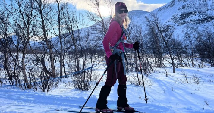 Vrouw skiet in besneeuwd landschap met bergen en bomen op de achtergrond.